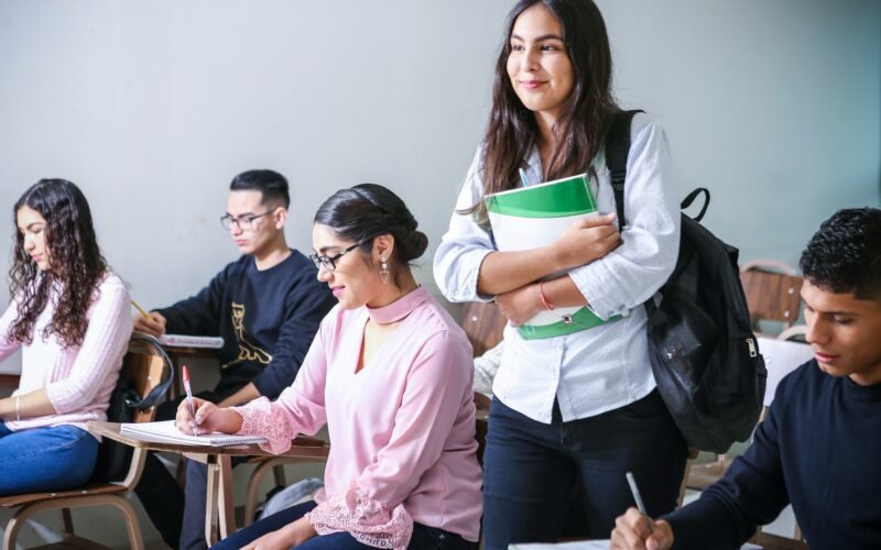 A realistic photograph titled javier-trueba-iQPr1XkF5F0-unsplash.jpg showing a diverse group of university students in a bright classroom setting. In the foreground, a smiling female student stands holding a green notebook and wearing a black backpack, while other students are seated at wooden desk chairs. To her left, a student in a pink blouse takes notes in a notebook, and a student in a black sweater is seated behind her. In the lower right corner, a male student focuses on writing at his desk.
